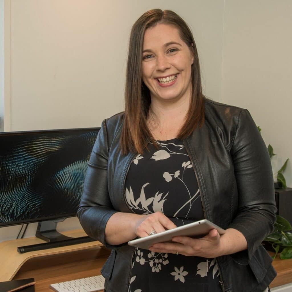 A person stands in an office holding a tablet, smiling. A computer monitor and keyboard are visible on the desk behind them.