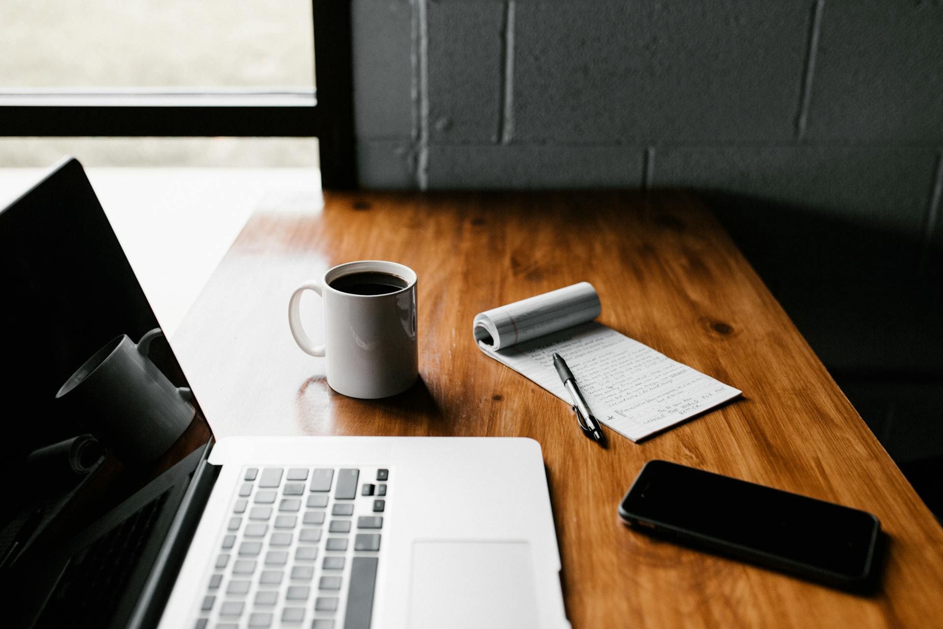 A laptop, coffee mug, notepad with pen, and smartphone on a wooden desk next to a window.