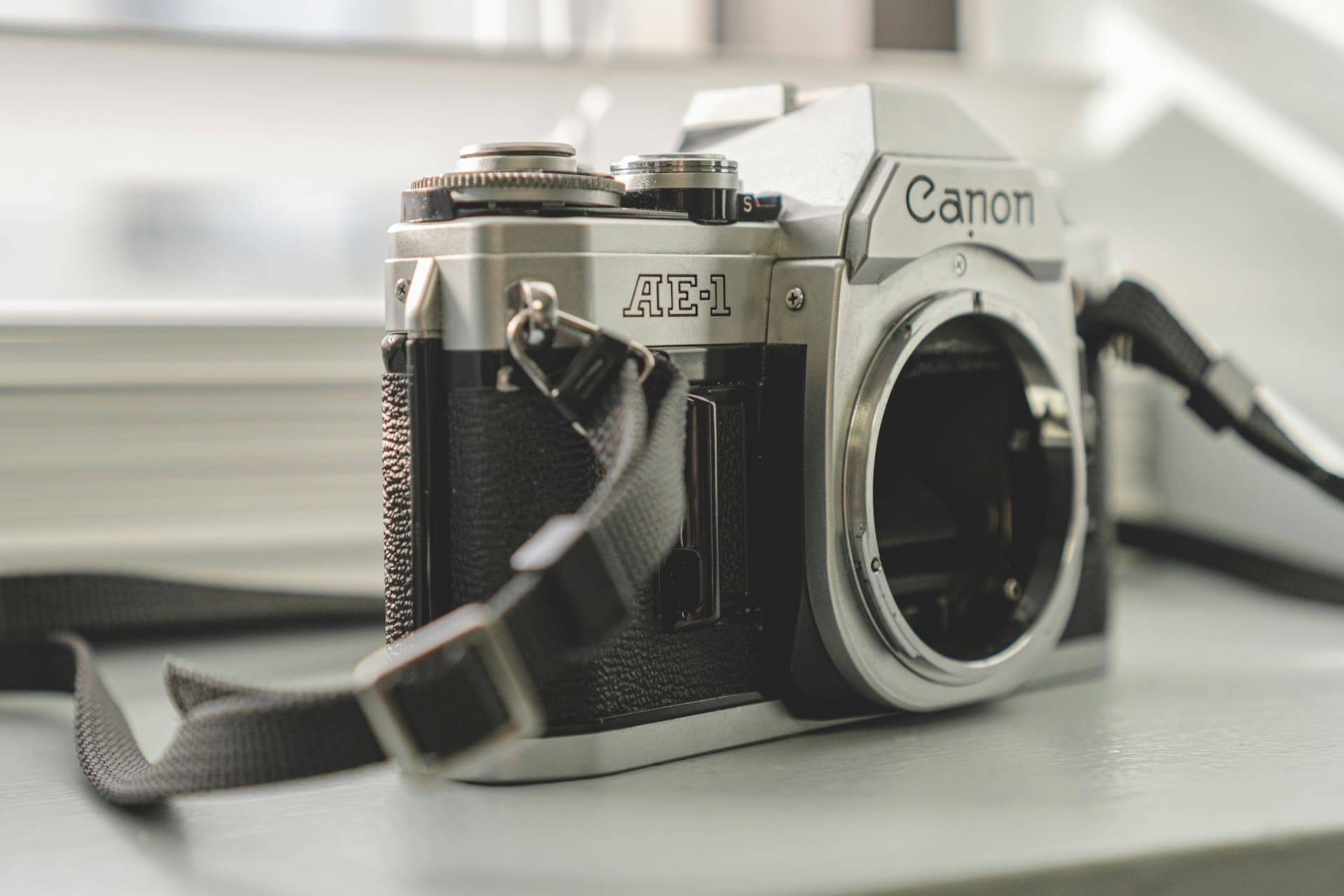 A Canon AE-1 35mm film camera with a black strap is placed on a gray surface, with its lens removed and a blurred background.