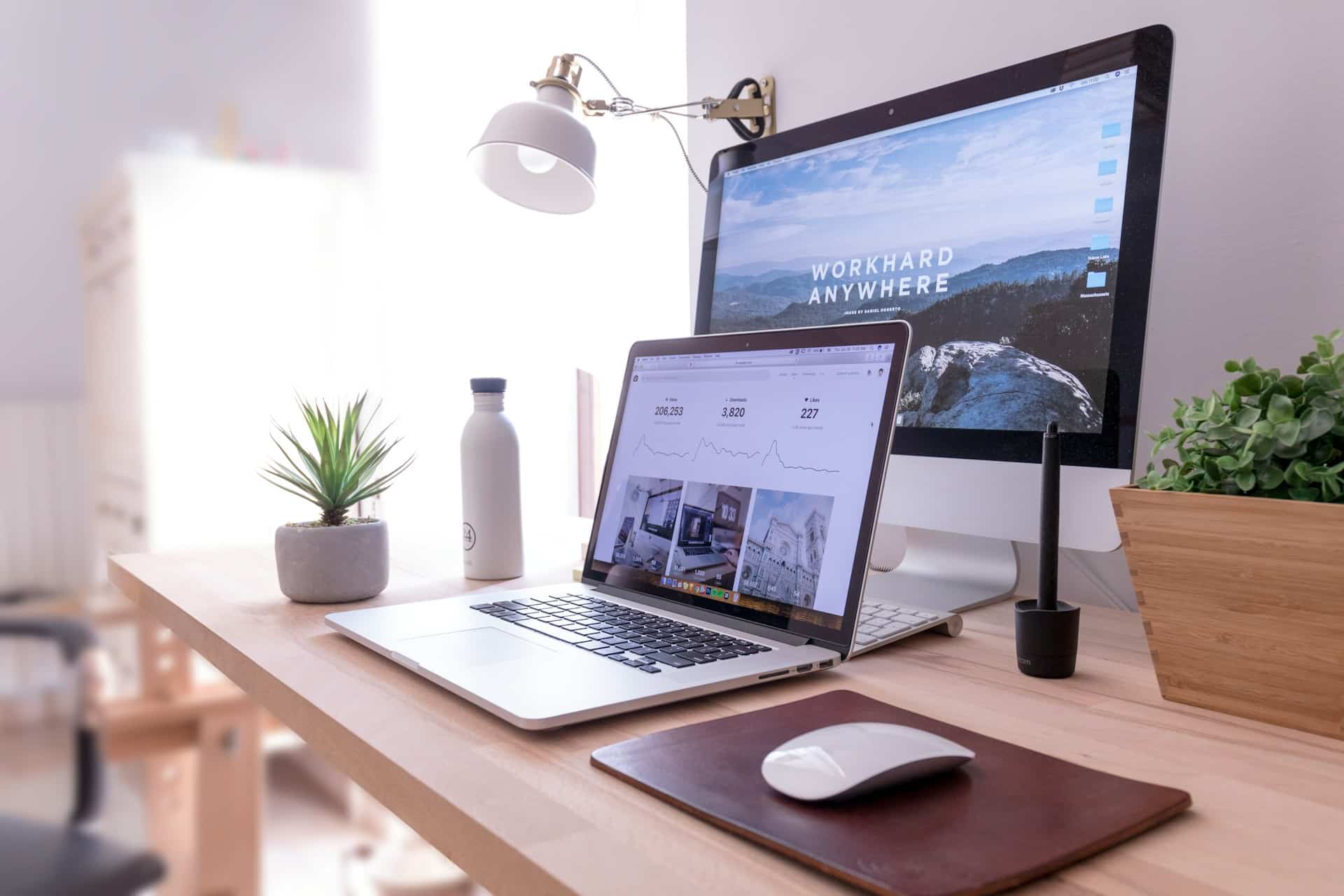 A tidy desk with a laptop, desktop monitor, keyboard, mouse, water bottle, potted plants, pen holder, and mousepad. The monitor displays "WORK HARD ANYWHERE" text.