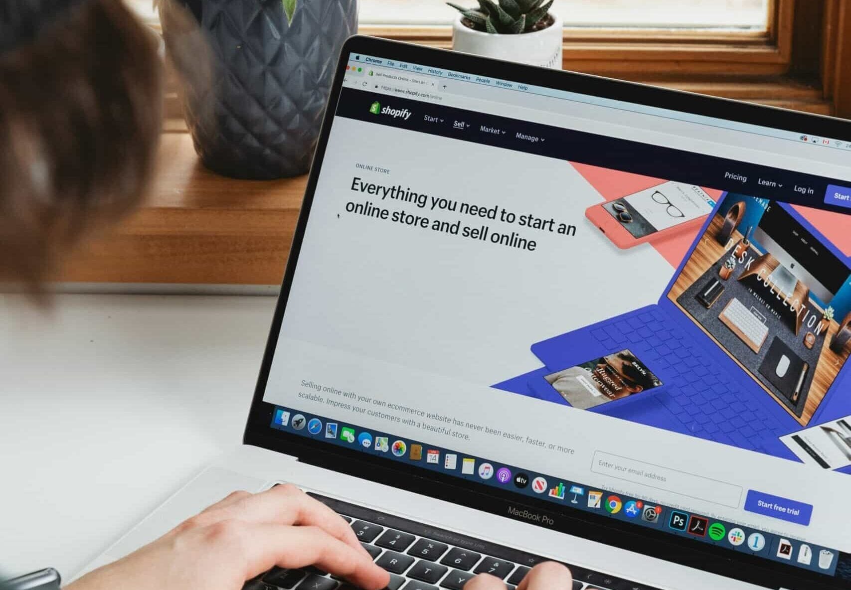 Person typing on a laptop displaying a Shopify webpage, with potted plants on the windowsill in the background.
